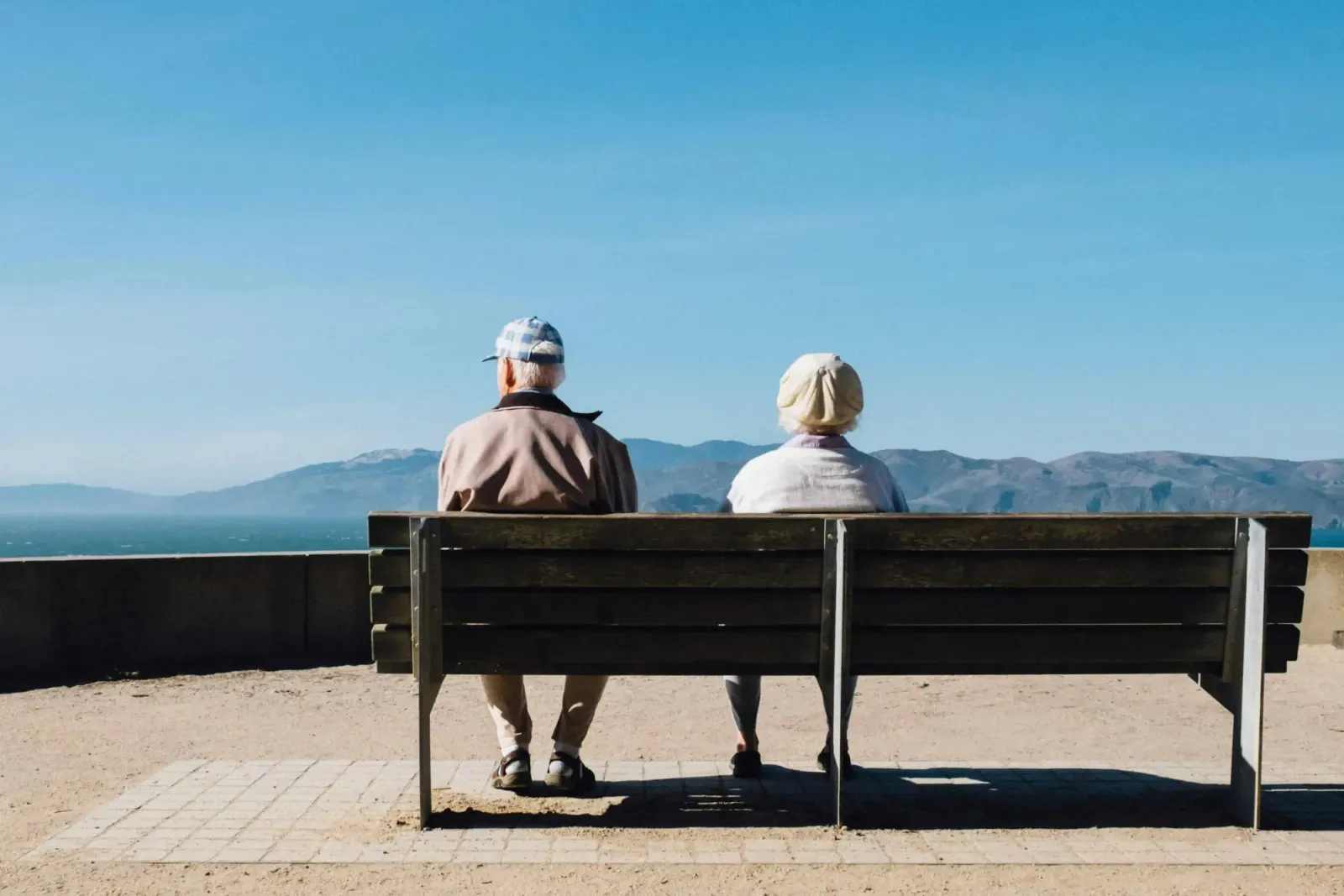 Elderly couple seen from behind sitting on a bench looking out over water and hills