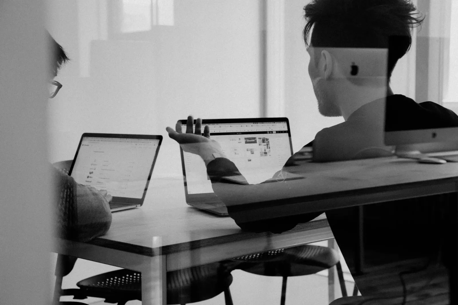 Two colleagues at a shared desk discussing work on their laptops in a black-and-white office photo.