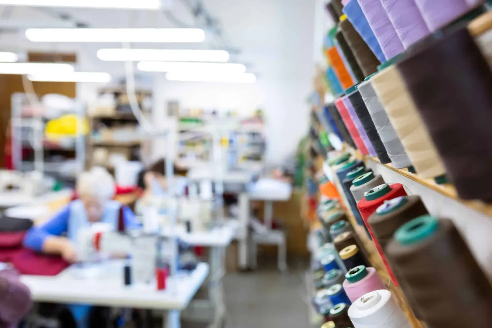 Apparel factory floor with sewing workers in the background and shelves of thread spools in the foreground